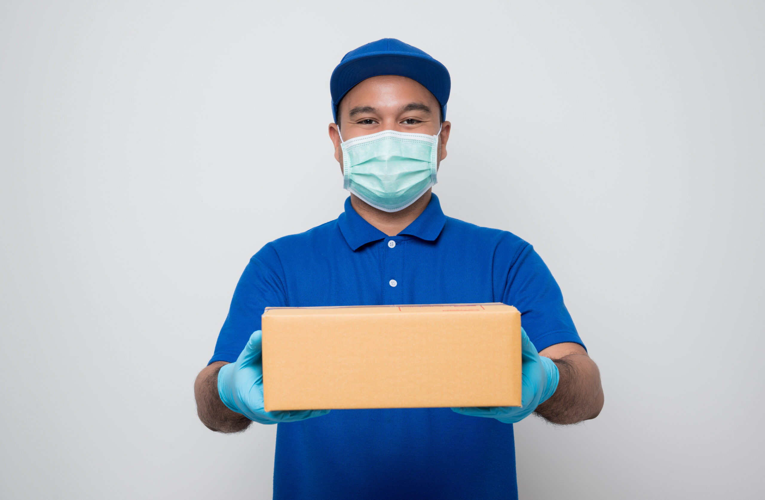 Young asian delivery man in blue uniform wearing protection mask and medical rubber gloves giving parcel cardboard to customer on isolated. Safety deliver concept.