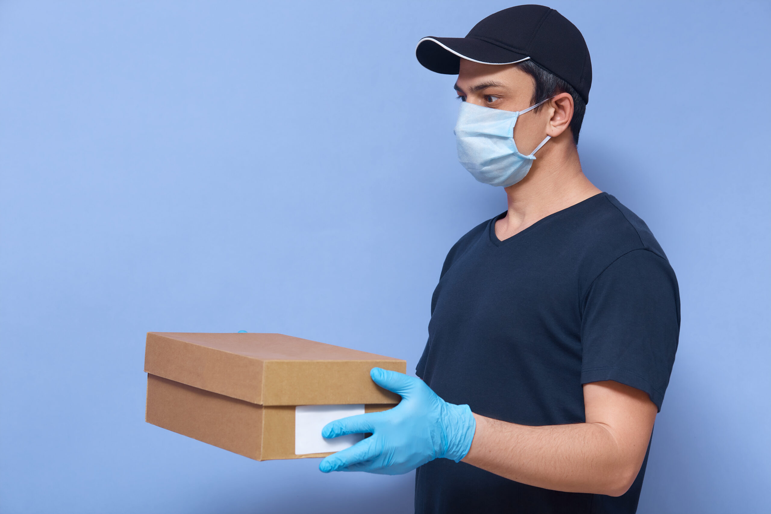 Indoor shot of young delivery man with carton box in hands, male wearing t shirt, cap and protective mask and latex gloves, meet his client to give order,posing isolated over blue studio background.